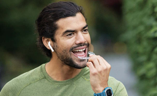 Man smiling while wearing earbuds and touching his face outdoors in casual green shirt.