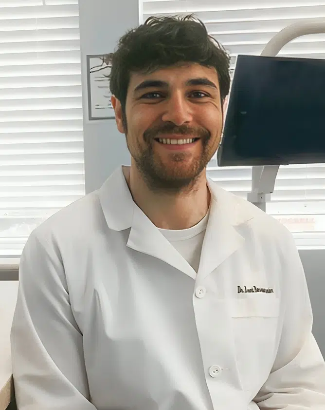 Man in white dental coat smiling in modern dental office setting.