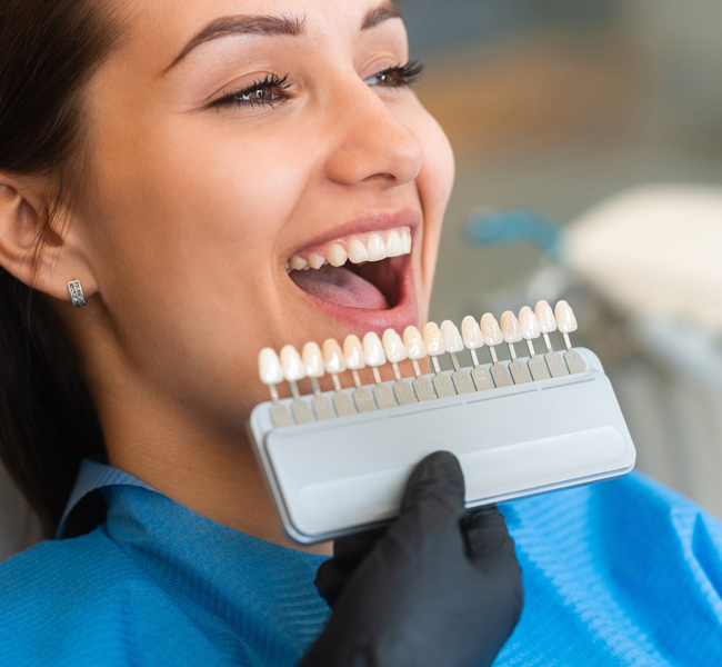 Woman holding tooth shade guide comparing dental veneers or whitening options with dentist.