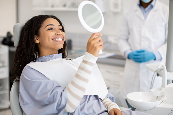 Patient examining mirror in dental office while dentist in background prepares examination.