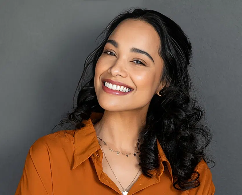 Smiling woman with long dark hair wearing orange shirt against gray background.