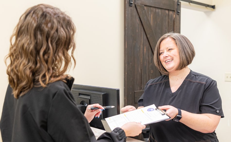 Two women in professional attire exchange documents in a dental office reception area.