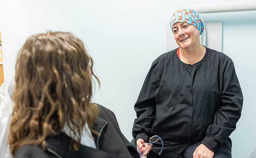 Female dental professional in surgical cap speaking with patient in clinical office setting.