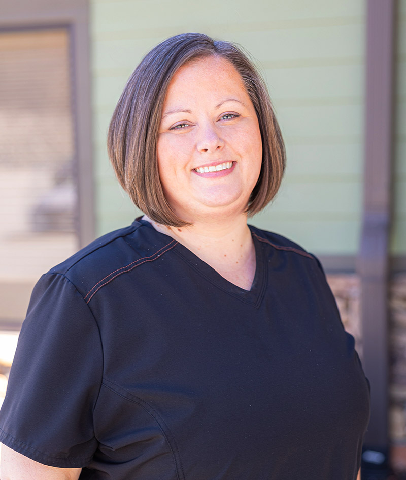 Professional headshot of woman in dark scrubs smiling at camera in medical office setting.