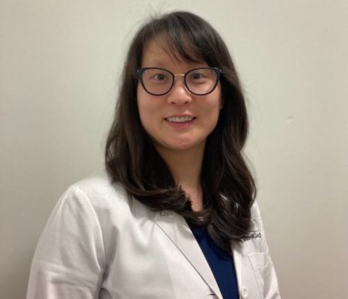 Professional headshot of a woman wearing glasses and a white coat in a medical office setting.